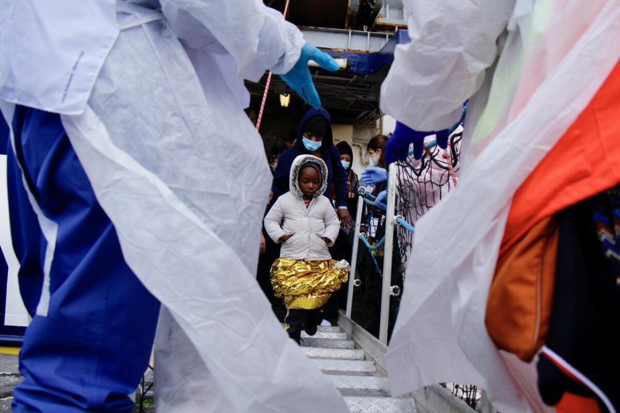 Kind in weißer Jacke und mit Rettungsdecke als Rock geht von der Humanity 1 an Land. Links und rechts sind die Schutzanzüge der wartenden Menschen am Hafen zu sehen.