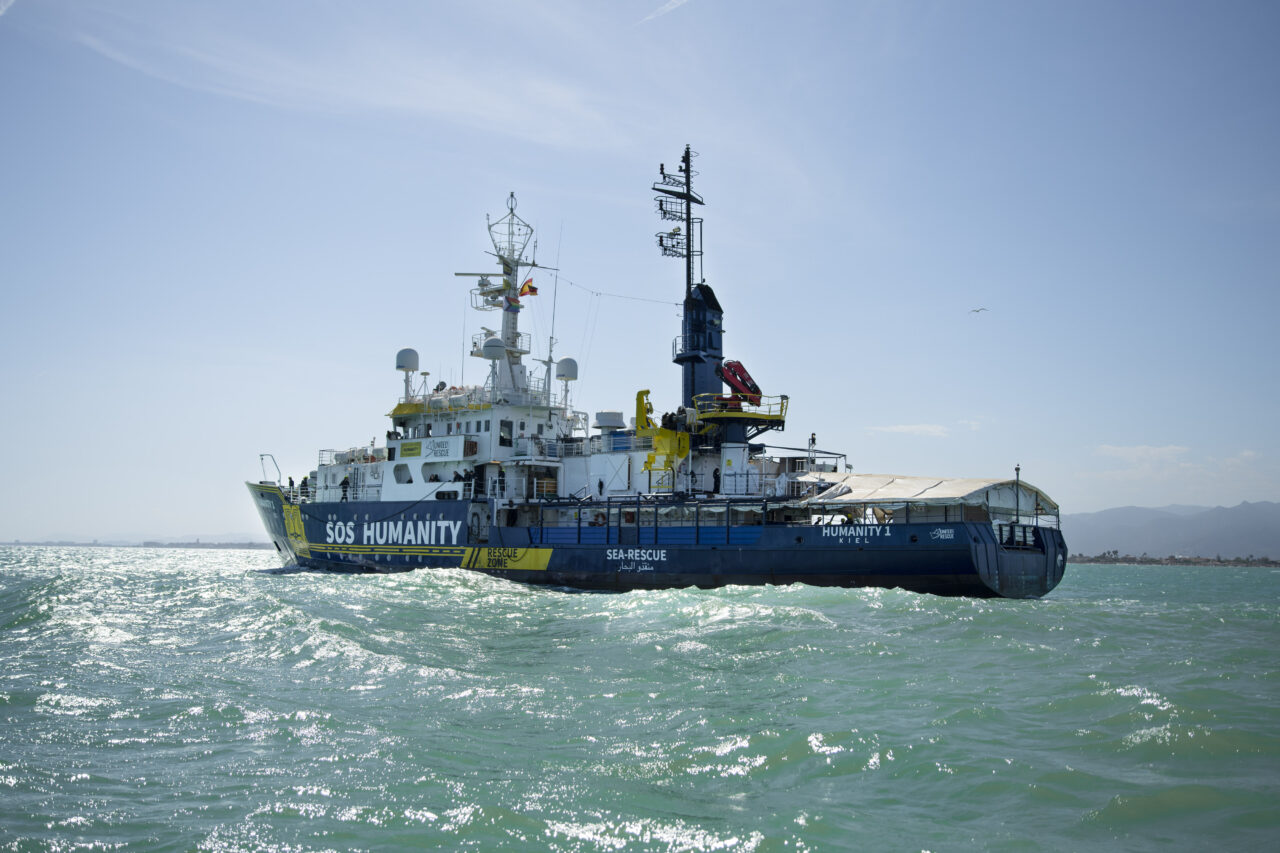 Humanity 1, das Rettungsschiff von SOS Humanity, auf türkisem Wasser (Mittelmeer) und mit blauem Himmel im Hintergrund.