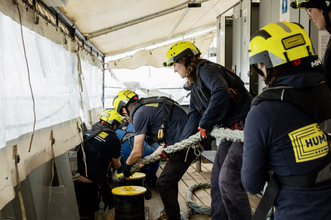 Crew an Bord der Humanity 1 zieht gemeinsam an einem Seil. Sie tragen gelbe Helme mit dem SOS Humanity Logo.