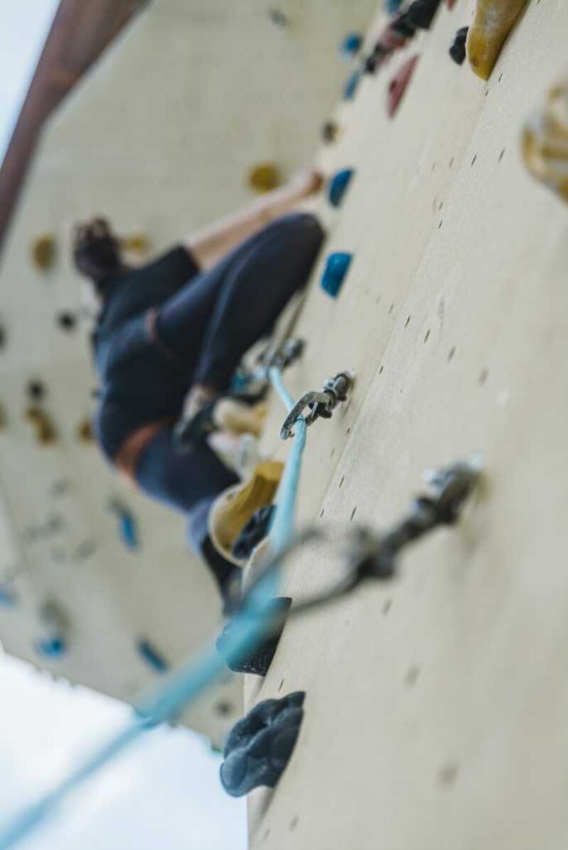 Kletterwand mit bunten Griffen und blauem Kletterseil und einer dunkel gekleideten Person, die gerade hochklettert.
