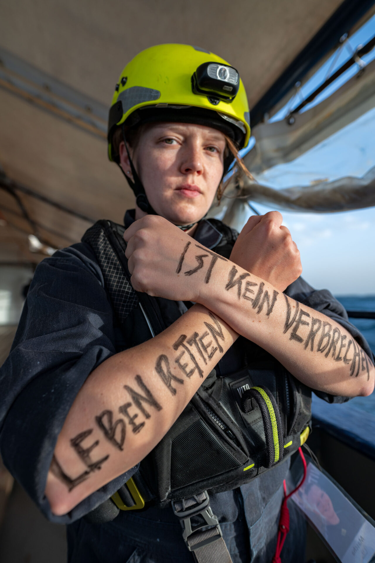 Crewmitglied mit gelben Helm auf dem Kopf, dem Schriftzug "Saving Lives is Not a Crime" auf den Armen, die vor der Brus gekreuzt sind an Bord der Humanity 1.