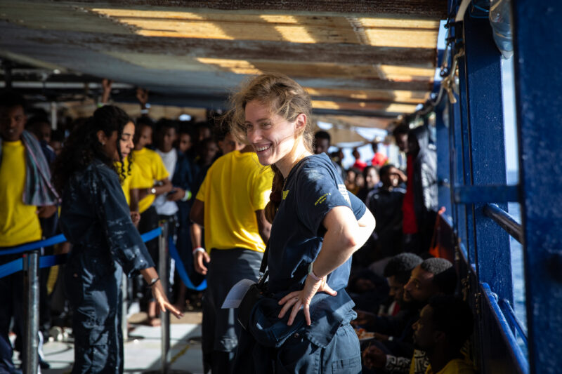 Crewmitglied lächelnd an Bord, Überlebende im Hintergrund