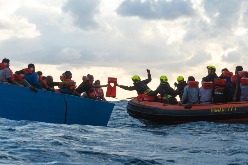 Scene of a rescue operation with the RHIB of SOS Humanity on the rights side and a refugee boat on the left side. One crew member is leaning over to the other boat to hand over life jackets.