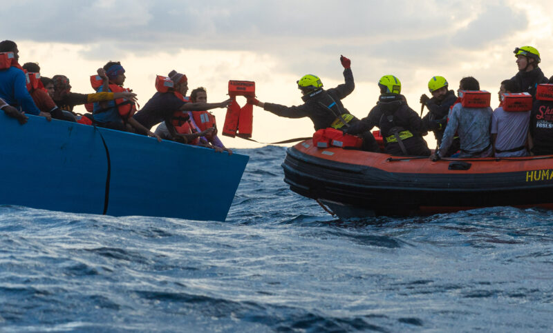 Scene of a rescue operation with the RHIB of SOS Humanity on the rights side and a refugee boat on the left side. One crew member is leaning over to the other boat to hand over life jackets.