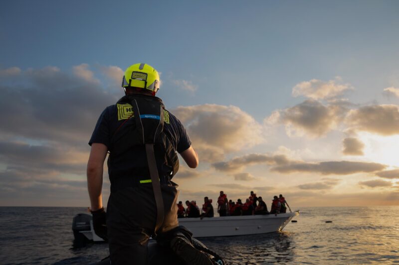 Foto von einem Crewmitglied von SOS Humanity mit gelbem Helm, das auf ein Flüchtlingsboot schaut