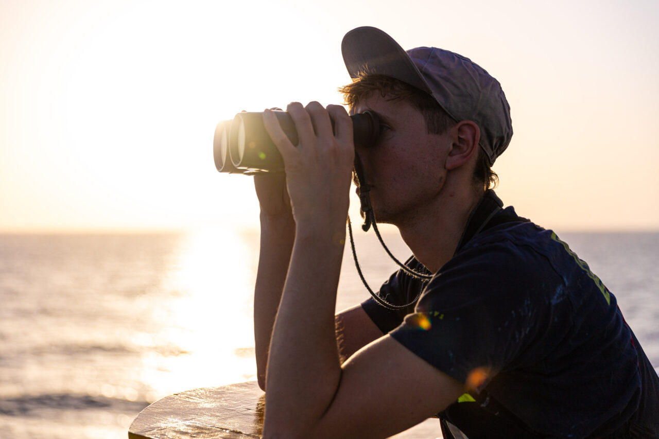 Ein Crewmitglied mit Kappe schaut durch ein Fernglas auf das zentrale Mittelmeer bei Sonnenuntergang