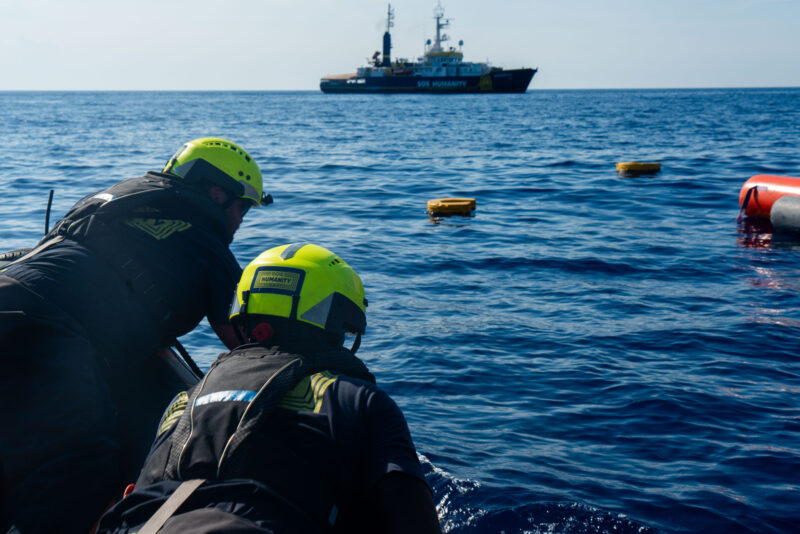 Zwei Crewmitglieder sind von hinten auf dem Foto zu sehen, während sie Rettungsringe ins Wasser werfen