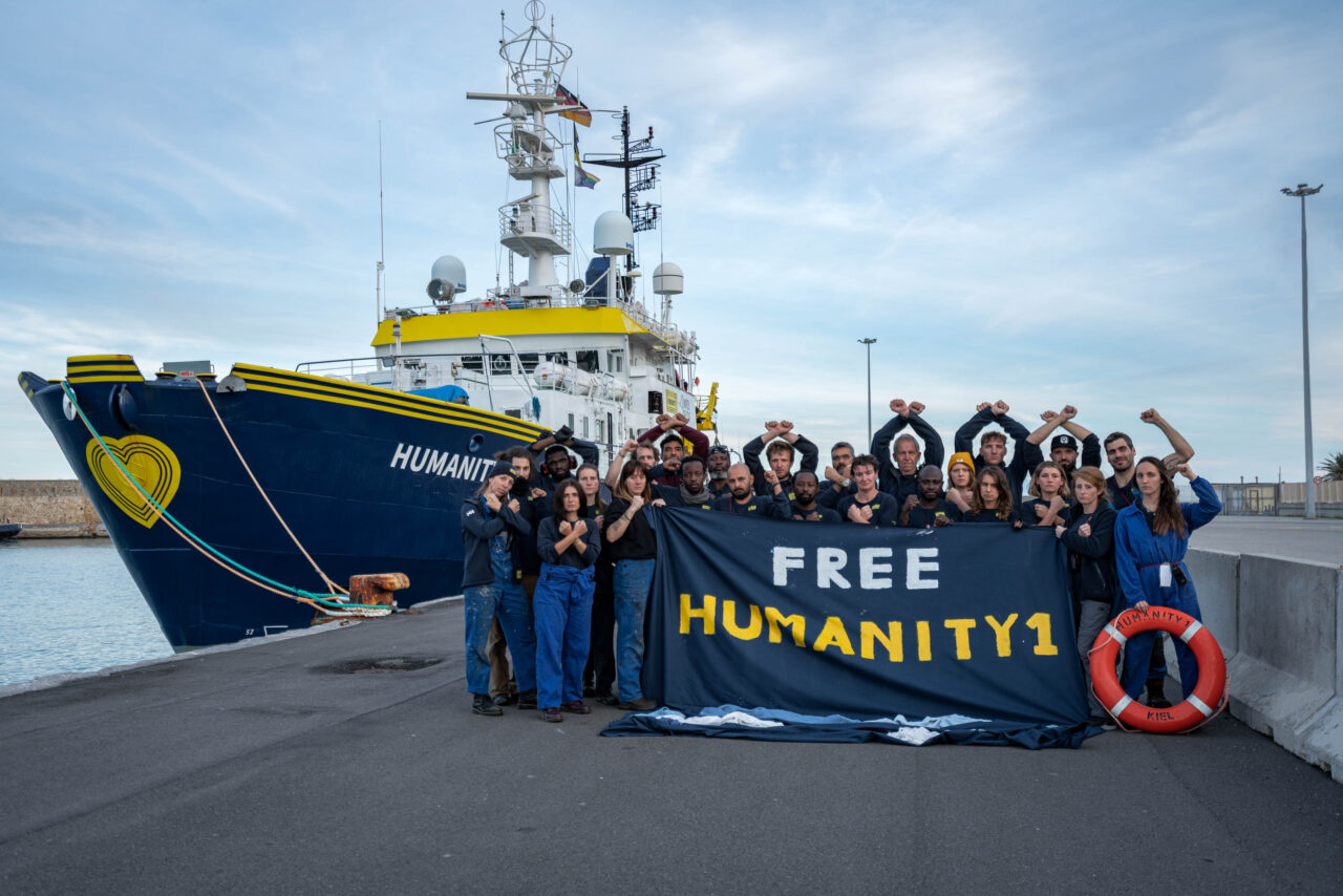 Die Crew von SOS Humanity steht vor dem Schiff Humanity 1 im Hafen und hält ein Banner mit der Aufschrift "Free Humanity 1"