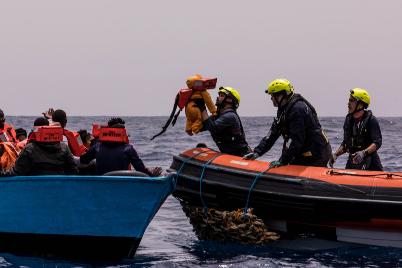 Das Bild zeigt eine Rettungsaktion von SOS Humanity, bei der ein Kleinkind von einem seeuntauglichen Boot in das Rettungsboot unserer Crew gehoben wird.