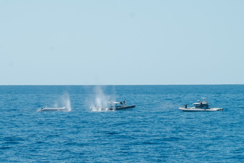 Das Bild zeigt ein Schlauchboot und zwei Boote der sogenannten libyschen Küstenwache, die auf dieses Schlauchboot schießen