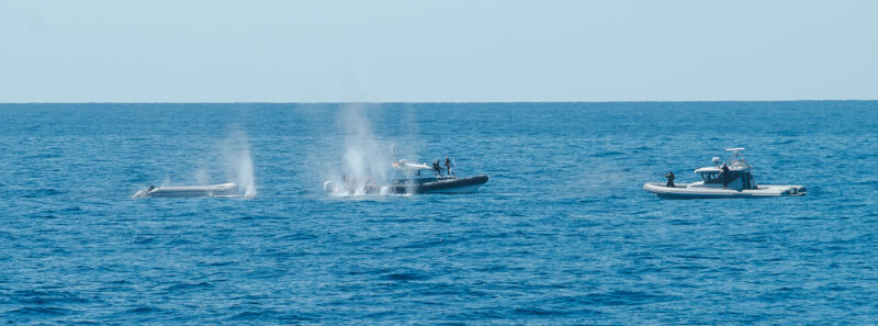 Das Bild zeigt ein Schlauchboot und zwei Boote der sogenannten libyschen Küstenwache, die auf dieses Schlauchboot schießen