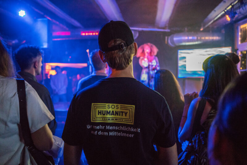 Ein Gast in einem SOS Humanity T-Shirt steht vor der Bühne im FluxBau.