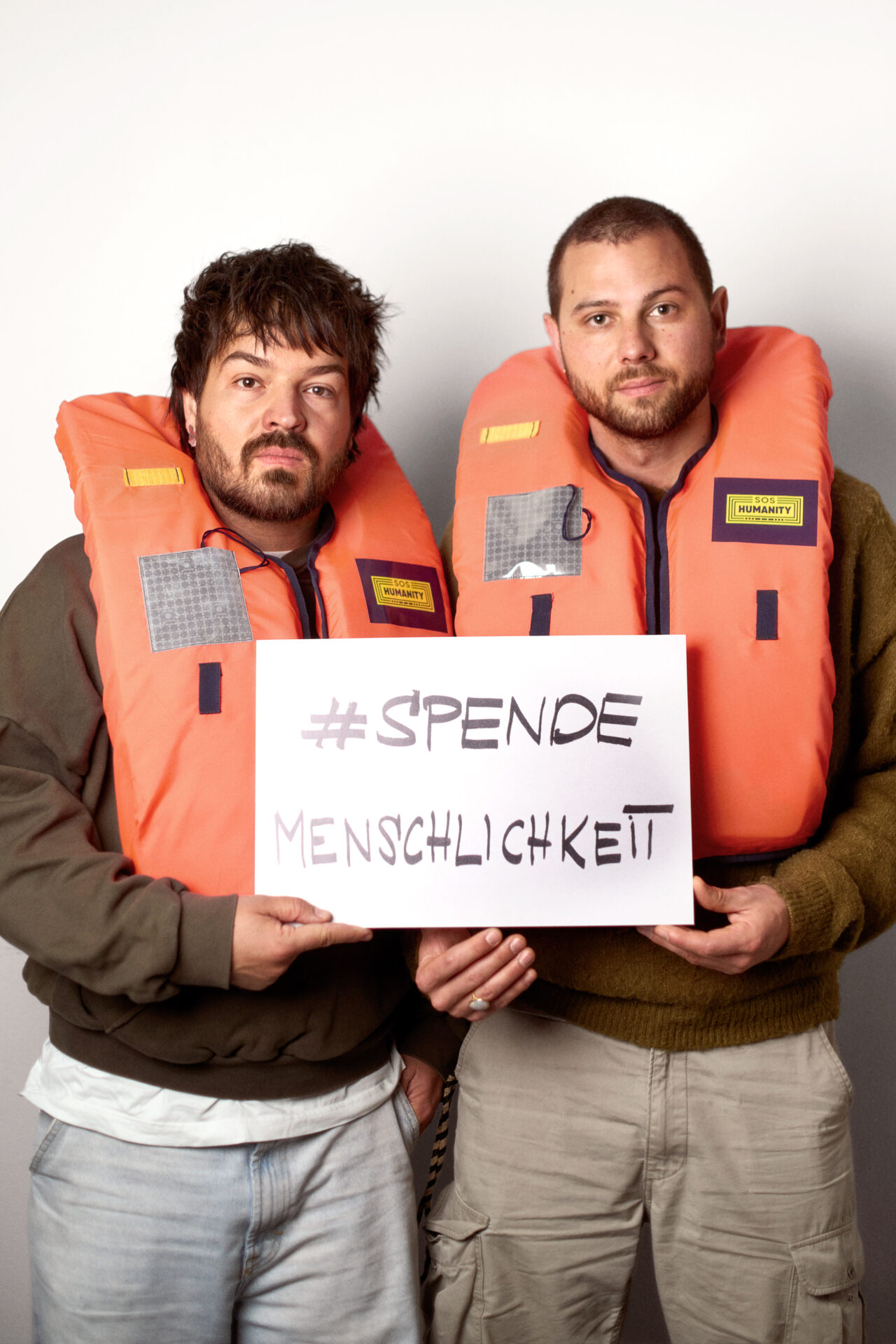 Zwei Männer von "Milky Chance" mit einer Rettungswesten und dem Schild 'SPENDEMENSCHLICHKEIT
